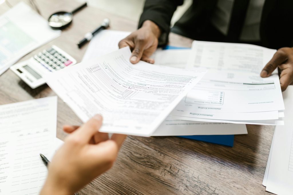 pexels-photo-7821684-7821684 Two professionals exchanging documents in an office setting, focusing on paperwork and data analysis.